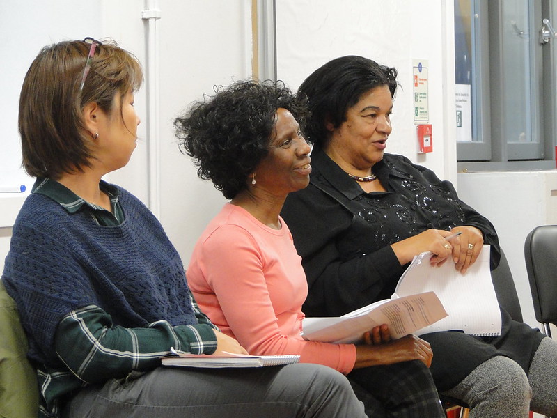 Three women sit on chairs talking at a community meeting
